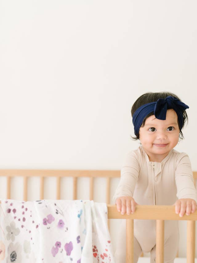 image of baby in a crib, organic cotton muslin swaddle blanket draped over the side of the crib. White blanket with floral print.