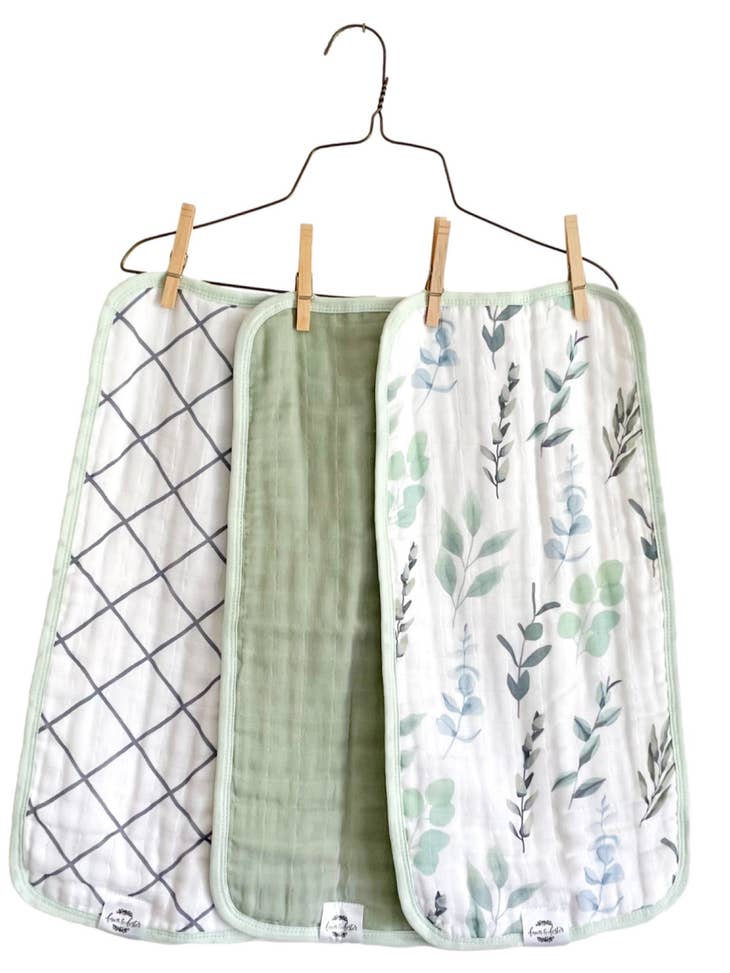Image of three organic cotton muslin burp cloths with different prints, with sage accent colors, shanging on hanger.