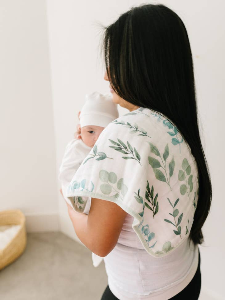 Image of woman holding an infant, whe has a burp cloth draped on her shoulder, it's white with sage green plant leaves on it. 