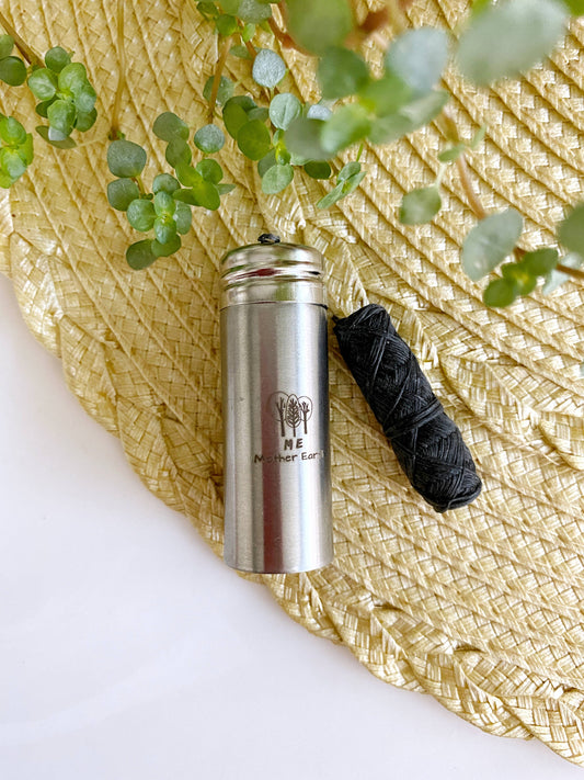 small stainless steel container with a bundle of black biodegradable dental floss laying next to it. Both items are laying on a woven place mat with greenery.